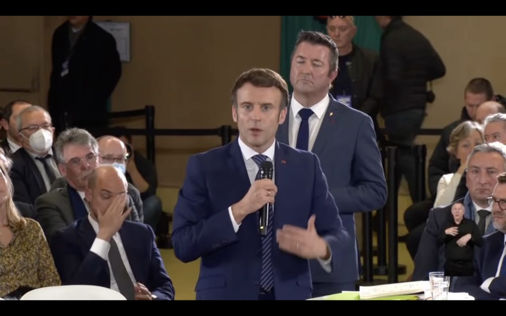 Capture d'écran: Emmanuel Macron intervient dans une réunion publique, il est debout avec un micro à la main, en arrière-plan, le public assis, et un médaillon LSF de petite taille, sans fond, est incrusté en bas à droite.