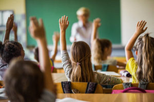 des élèves dans une salle de classe, de dos, lèvent la main, une enseignante en arrière-plan fait face à la classe