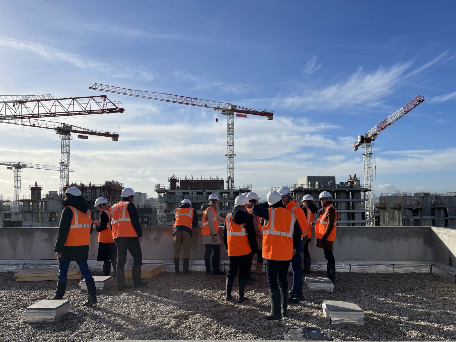 photo d’un chantier avec des grues, des immeubles en construction et des personnes portant un casque de chantier et un gilet orange fluo