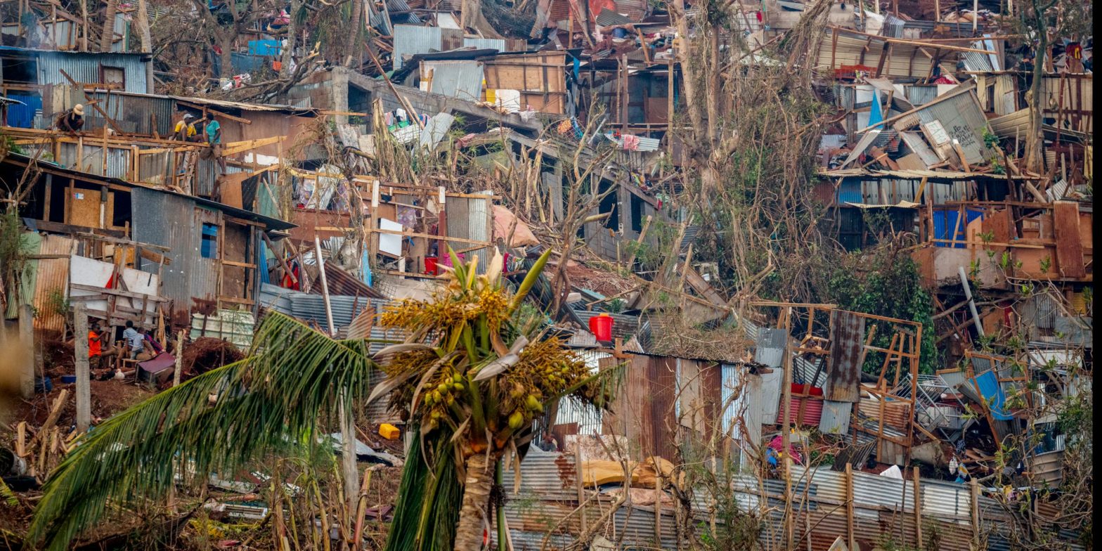 Mayotte après le passage du cyclone Chido. Sur cette image, un quartier défavorisé construit à flanc de colline, constitué de nombreuses habitations faites de tôle, de bois et de matériaux de récupération. La scène montre des dégâts importants : de nombreuses maisons sont gravement endommagées, certaines se sont effondrées, d’autres n’ont plus de toit ou de murs. Des arbres sont tordus ou arrachés, des branches et des débris jonchent le sol. Au milieu de ces ruines, quelques personnes tentent de réparer ou de fouiller ce qu’il reste de leurs maisons. L’ensemble illustre la précarité et les difficultés que rencontrent les habitants après le passage du cyclone.
